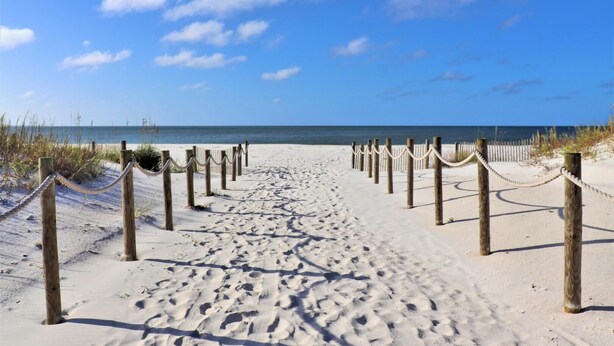 Un sentier de sable qui longe la plage et mène à l’océan, bordé de poteaux en bois et de barrières faites de cordes.