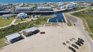 Vue aérienne d’une station balnéaire dotée d’un port de plaisance, d’un parking sablonneux et de bâtiments aux grandes fenêtres donnant sur l’eau.