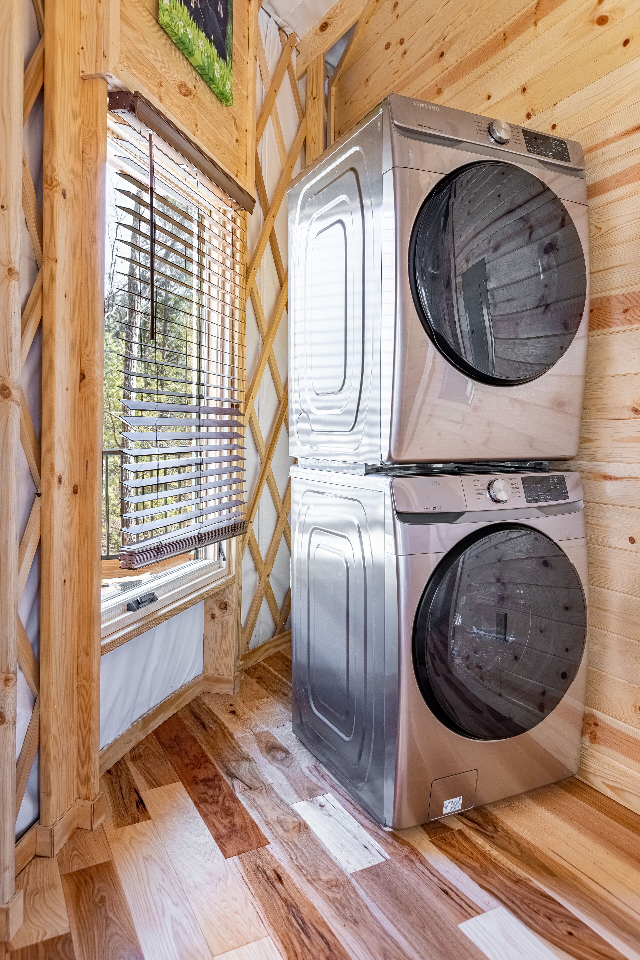 A compact room with two stacked stainless steel front-loading washing machines, a wooden lattice window, and a wooden floor.