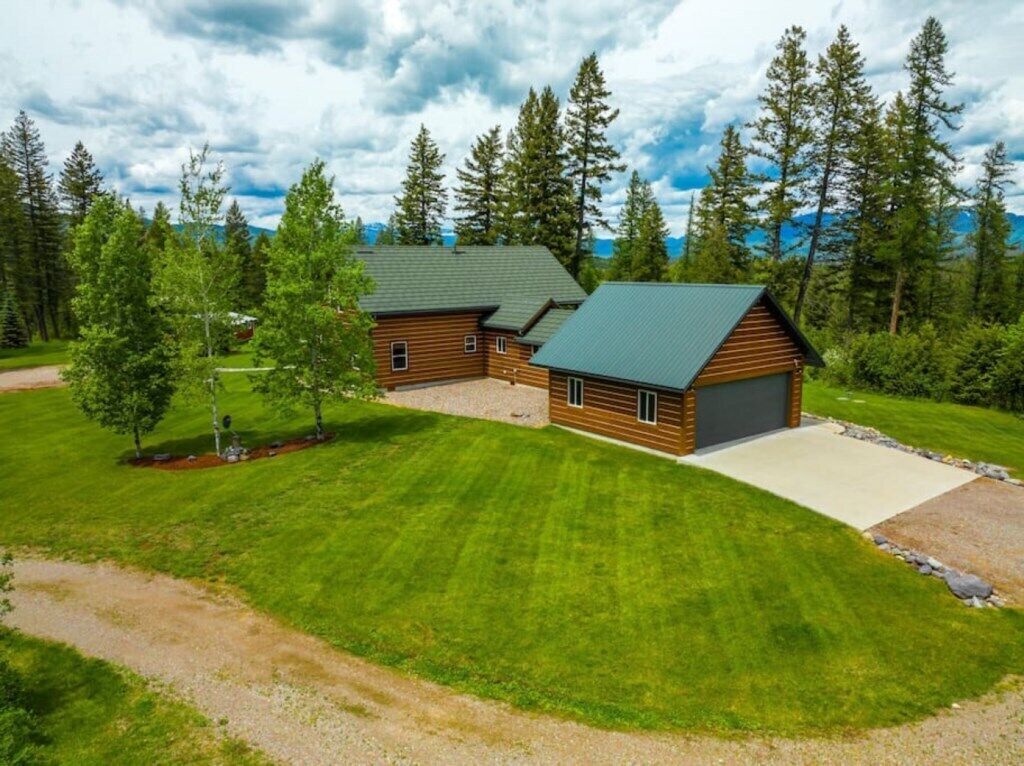 A two-story log cabin with a green roof, surrounded by a well-maintained lawn and tall trees.