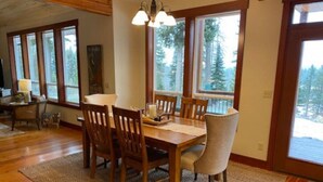 A dining area with a wooden table and chairs, a woven rug, and large windows with a view of trees.