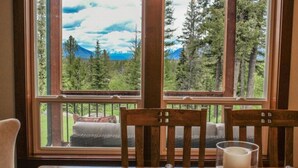 A room with a large window overlooking a forest and mountains. There are two wooden chairs and a table with a candle.
