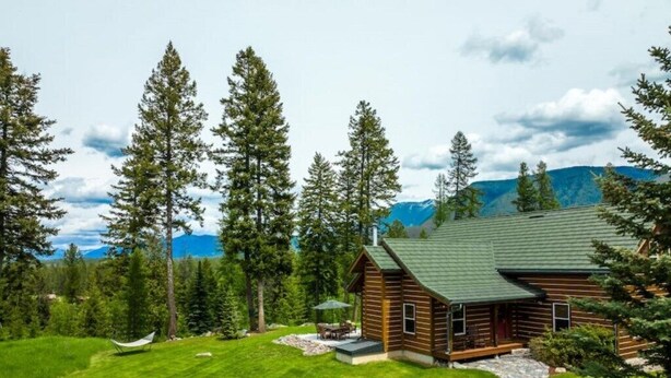 A log cabin with a stone pathway leading to a patio, surrounded by a well-maintained lawn and tall trees.