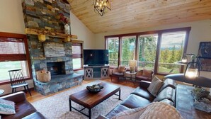 A cozy living room with a stone fireplace, leather sofas, a television, and large windows offering a view of the outdoors.