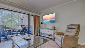 A living room with a glass coffee table, a leather armchair, a TV mounted on the wall, and a view of the outdoors through large windows. - Romantic Getaway II w/ WiFi in Oceanfront Gated Resort - 1 Bedroom Lodge (Myrtle Beach)