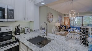 A modern kitchen with white cabinets, a built-in microwave, a stove, and a sink. There is a dining area with a table and chairs, and a large window providing a view of the outdoors. - Romantic Getaway II w/ WiFi in Oceanfront Gated Resort - 1 Bedroom Lodge (Myrtle Beach)