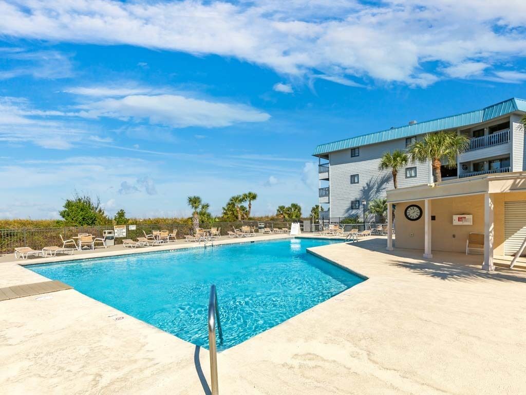Una piscina con sillones de descanso, un edificio con techo azul y palmeras al fondo.