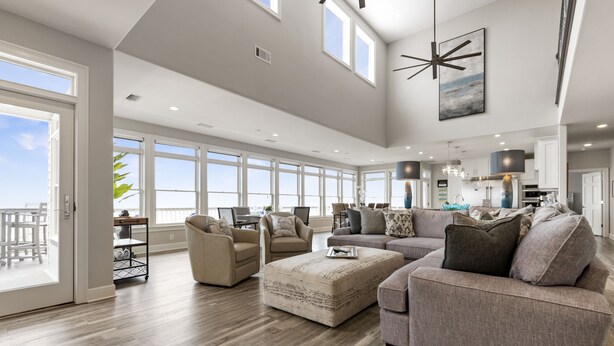 A spacious living room with a large window, a sofa, a coffee table, and a dining area visible through the glass doors. - Endless Summer-Gulf Front custom home for 20+.  Pup friendly! (Navarre)