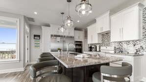 A modern kitchen with white cabinets, a large island with bar stools, stainless steel appliances, and a view to the outside. - Endless Summer-Gulf Front custom home for 20+.  Pup friendly! (Navarre)