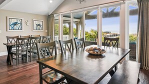 Un coin repas avec une table et des chaises en bois, une grande fenêtre donnant sur un paysage verdoyant et des œuvres d’art murales sur le thème de la marine.