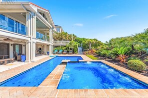 A swimming pool with a two-tiered deck, surrounded by lush greenery and a multi-story building with large windows.