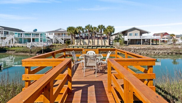 A wooden deck with a table and chairs, overlooking a calm body of water and beachfront properties.