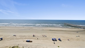 A wide sandy beach with a pier extending into the water, several umbrellas, and people relaxing. - Sea Pointe 706 - 1 Bed/1 Bath Ocean Front Condo - Indoor Pool & Hot Tub (North Myrtle Beach)