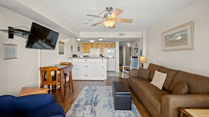 A living room with a brown sofa, a blue armchair, a wooden coffee table, and a TV mounted on the wall. - Sea Pointe 706 - 1 Bed/1 Bath Ocean Front Condo - Indoor Pool & Hot Tub (North Myrtle Beach)