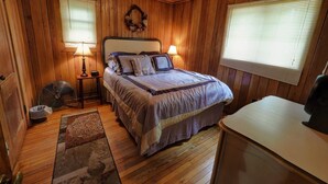 Une chambre à coucher aux murs lambrissés, avec un lit, deux lampes de chevet, un ventilateur et une commode.