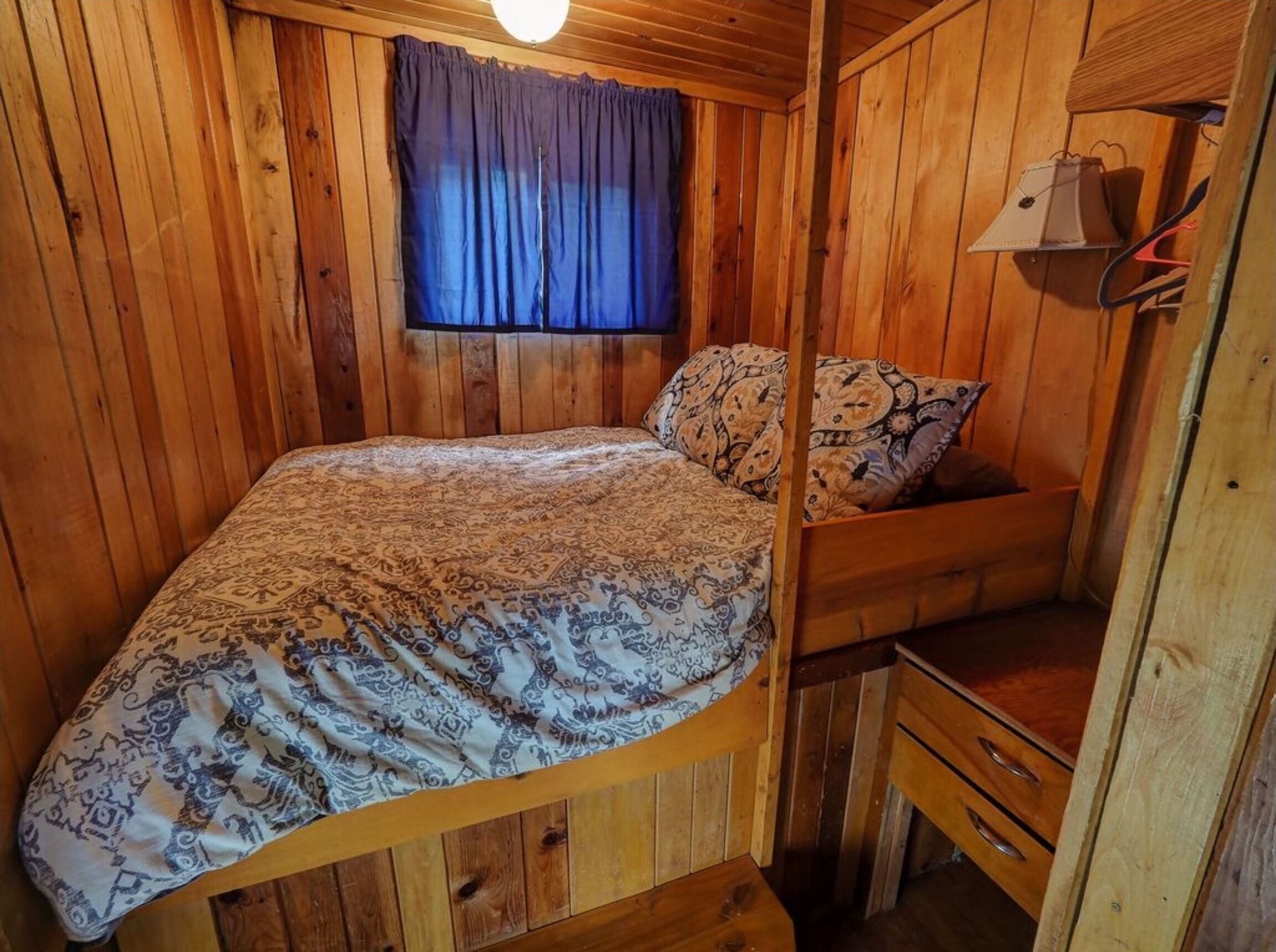 A wooden cabin room with a bed, a window with curtains, a small desk, and a hanging lamp.
