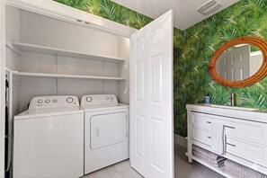 A laundry room with a washer and dryer, a white vanity with a mirror, and a door leading to another room.