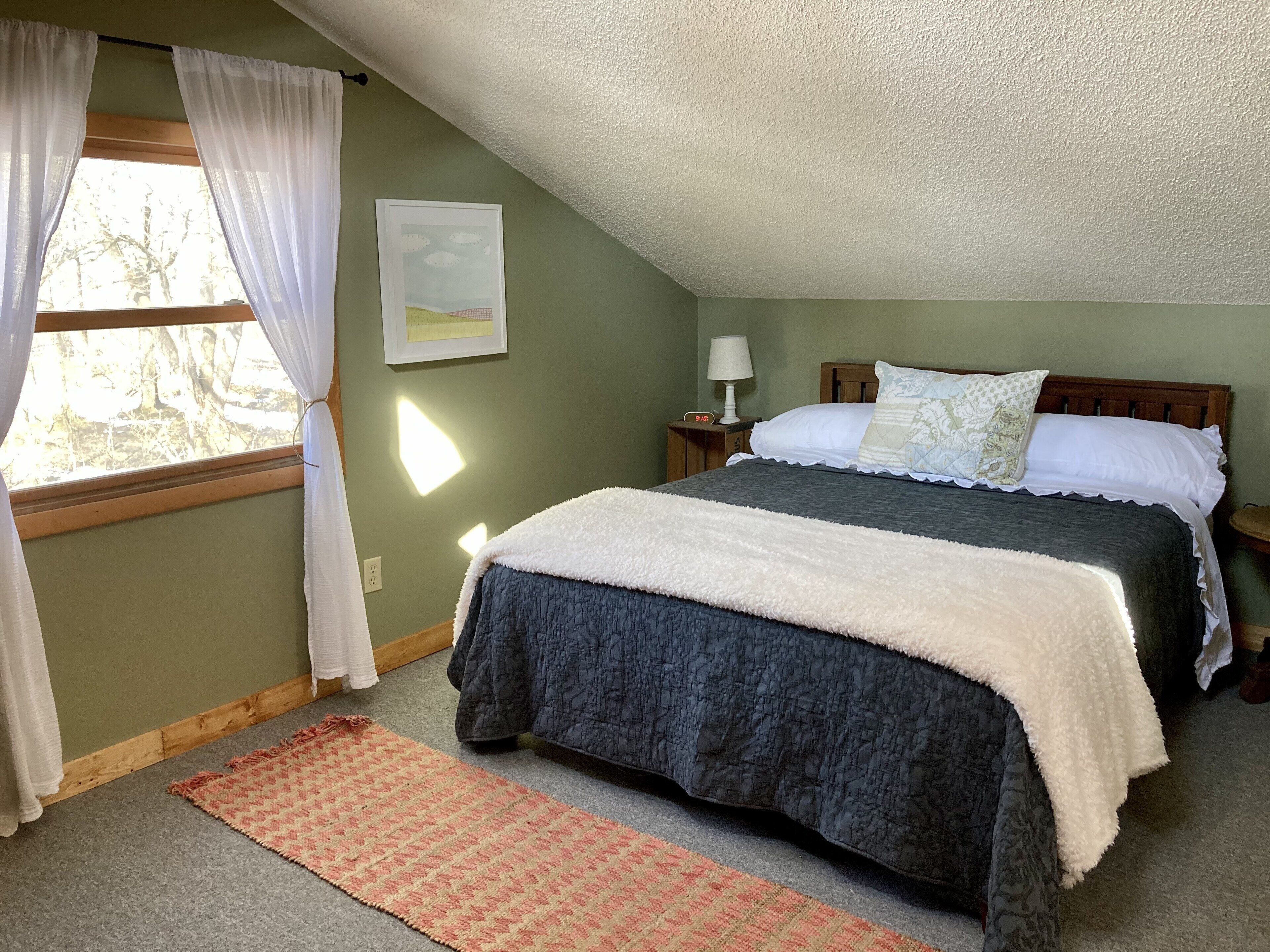 Un dormitorio con cama, mesita de noche, lámpara, ventana con cortinas e imagen colgada en la pared.