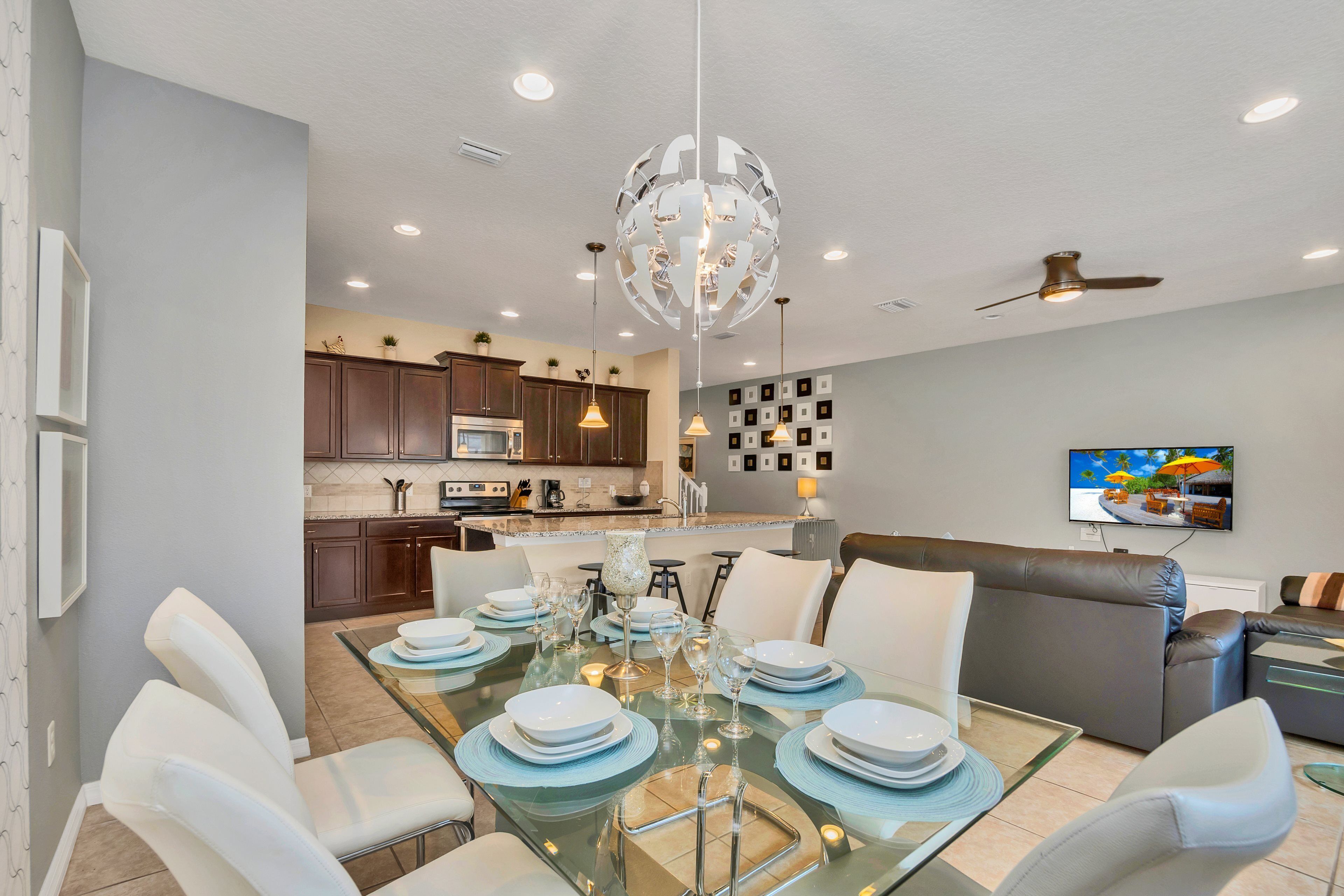 A modern dining area with a glass table set for six, surrounded by white chairs. A kitchen with dark cabinets and a central island is visible in the background.