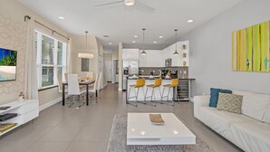 A modern living room with a white sofa, a coffee table, and a dining area with a white table and chairs. A kitchen with white cabinets and a bar area is visible in the background.