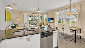 A modern kitchen with a dishwasher, dining area with white chairs, and a large window with blinds.