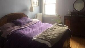 Een slaapkamer met een bed, nachtkastje, dressoir, raam en spiegel.