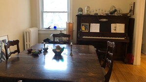 Een eetkamer met een houten tafel, stoelen, een piano en een raam met uitzicht op bomen.