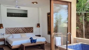 A bedroom with a bed, bedside tables, a nightstand, a wooden headboard, a ceiling fan, a wall-mounted light, a large window, and a bathtub. - Lazuli Playa - Beachfront Loft (Santa Maria Tonameca)