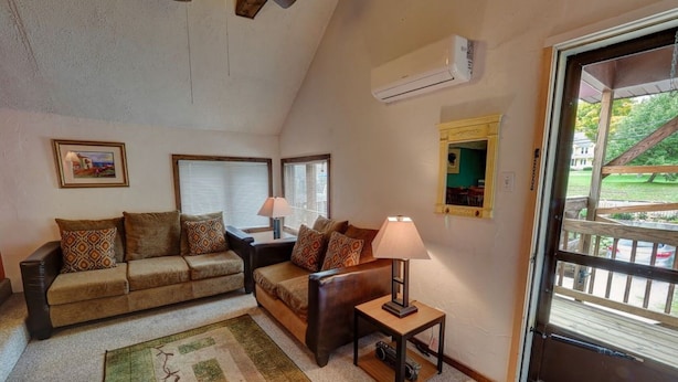 A cozy living room with a sofa, a smaller couch, a coffee table, and a lamp. There is a window with blinds, an air conditioning unit, and a door leading outside. - Upper Level Loft in Downtown Suttons Bay Across the Street From Beach and Marina (Suttons Bay)