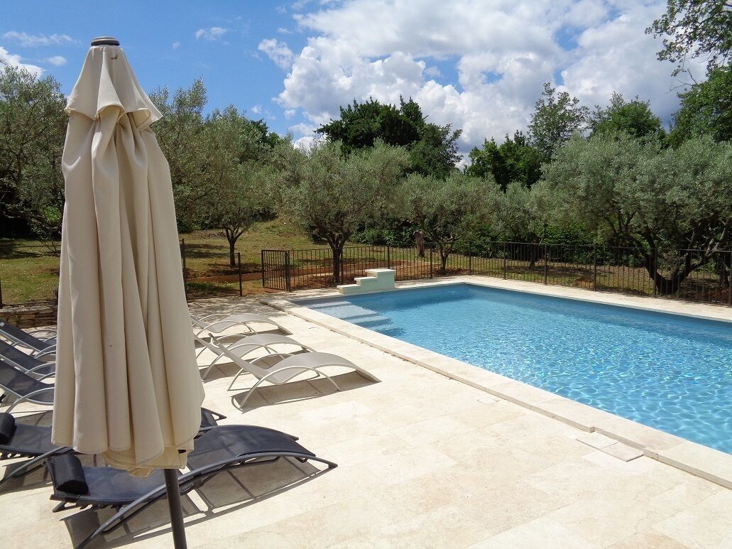 Una piscina con sillas de descanso y una sombrilla, rodeada de árboles y una valla.