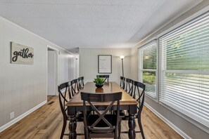 A dining room with a wooden table and chairs, a framed sign on the wall, and a window with blinds.