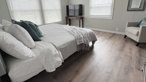 Un dormitorio con una cama grande, dos ventanas con persianas, un televisor sobre un mueble, una silla y un espejo en la pared.