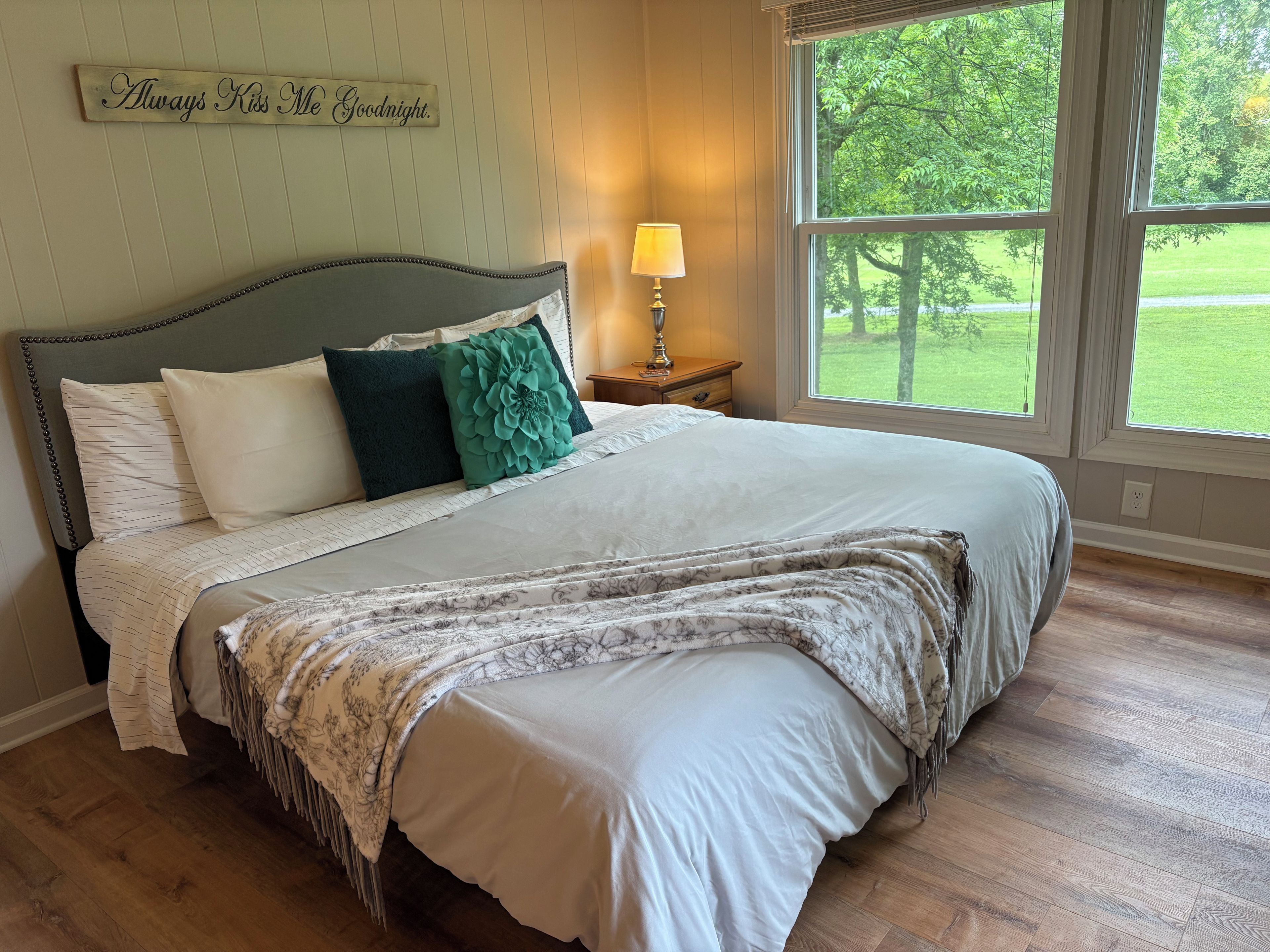 A neatly made bed with a floral throw blanket, a bedside lamp, and a view of a green landscape through a large window.