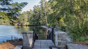 Ein Betonweg führt zu einem verschlossenen Eingangstor am See, umgeben von dichtem Baumbestand.