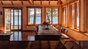 Ein geräumiger Speisesaal mit Holztisch und -stühlen, großen Fenstern mit Seeblick und einer hohen Decke mit sichtbaren Balken.