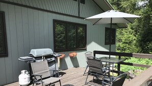 Een terras buiten met een barbecue, stoelen, een tafel en een parasol.