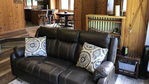 Een gezellige woonkamer met een leren bank, decoratieve kussens en een klein houten bijzettafeltje. Op de achtergrond is een eethoek met een ronde tafel en stoelen zichtbaar.