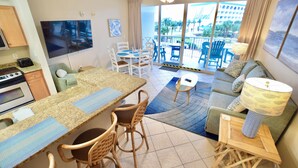 A compact hotel room with a kitchenette, a dining area with a table and chairs, a sofa, and a balcony with a view of a building and greenery. - Destin West Gulfside #309 ~Free Beach Service! Great View! Pools + Lazy River! (Fort Walton Beach)