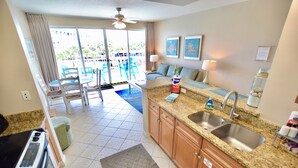 A kitchen with granite countertops, a double sink, and wooden cabinets. There is a dining area with blue chairs and a table, and a balcony with a view of the pool and palm trees. - Destin West Gulfside #309 ~Free Beach Service! Great View! Pools + Lazy River! (Fort Walton Beach)