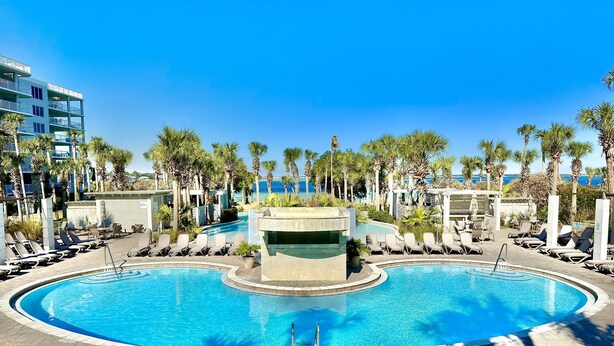 A pool area with lounge chairs, palm trees, and a view of the sea. - Destin West Gulfside #309 ~Free Beach Service! Great View! Pools + Lazy River! (Fort Walton Beach)