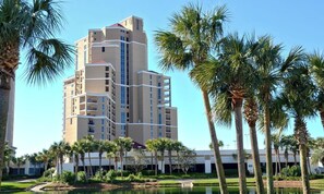 Un hotel a più piani con una torre centrale, circondato da palme e uno stagno.