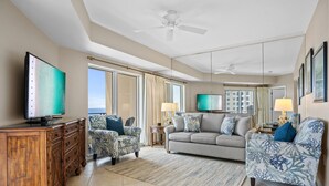 Ein geräumiges Wohnzimmer mit einem großen Fernseher, zwei gemusterten Sesseln, einem grauen Sofa und einer Holzkommode.