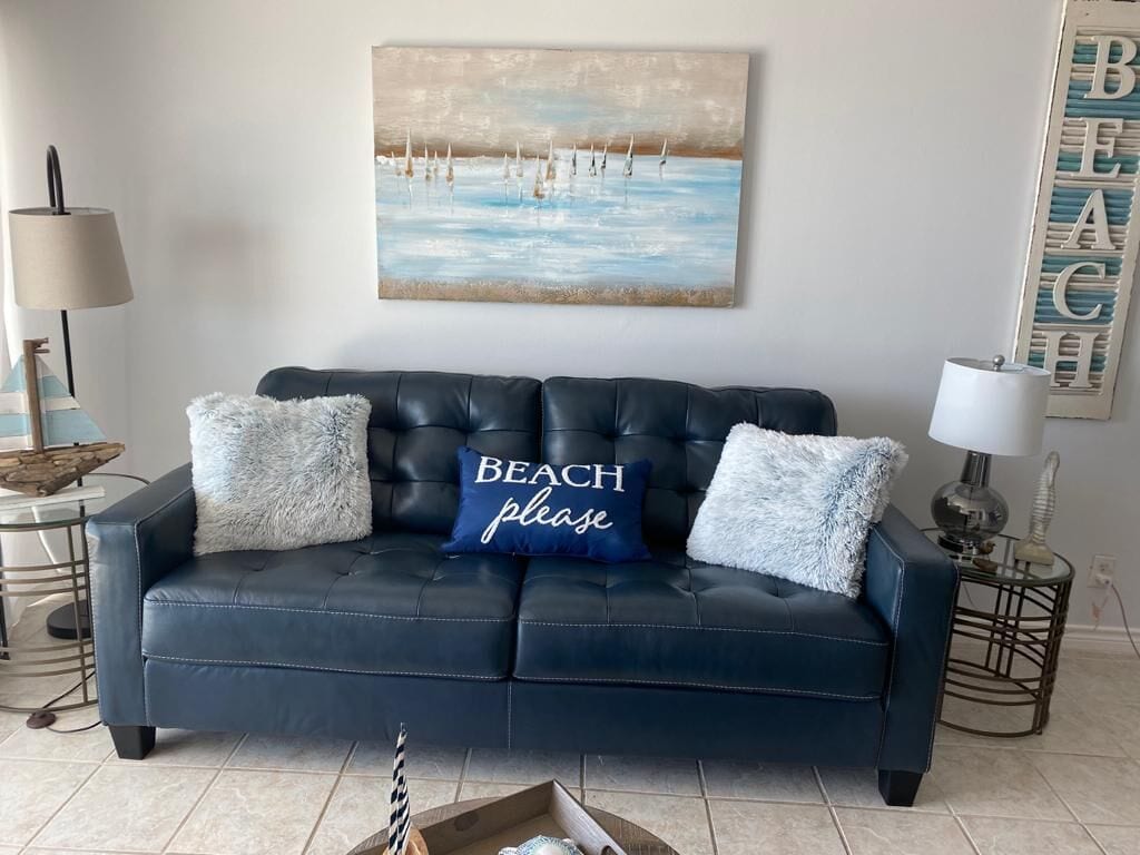 A dark blue leather sofa with two fluffy pillows, one with "BEACH please" text, a painting of sailboats on the wall, and a "BEACH" sign on the wall.
