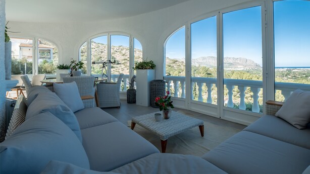 A spacious living area with a large sofa set, a small table with a vase, and a balcony with a view of the landscape.