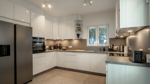 A modern kitchen with white cabinets, a stainless steel refrigerator, a built-in oven, and a window providing natural light.