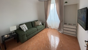 A living room with a green sofa, a small table with a lamp, a TV mounted on the wall, and a window with sheer curtains. - Appartamento Stazione Termini A/C WI FI (Roma)