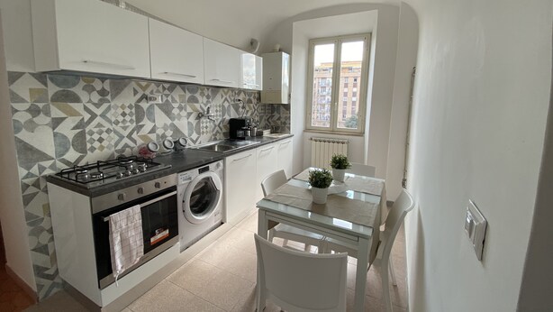 A compact kitchen with a gas stove, washing machine, and a dining area with a table and chairs. - Appartamento Stazione Termini A/C WI FI (Roma)