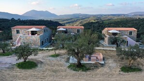 Un conjunto de edificios de piedra con techos de tejas rojas, rodeado de olivos y un paisaje montañoso al fondo.