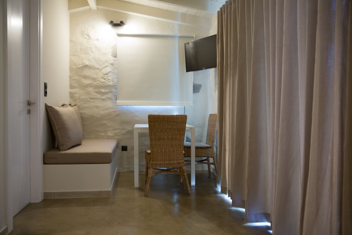 Una habitación pequeña y acogedora con una pared blanca, un televisor de pantalla plana montado sobre una mesa blanca y una silla de mimbre.