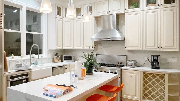 A modern kitchen with white cabinets, a stainless steel range hood, a large island with bar stools, and pendant lighting.